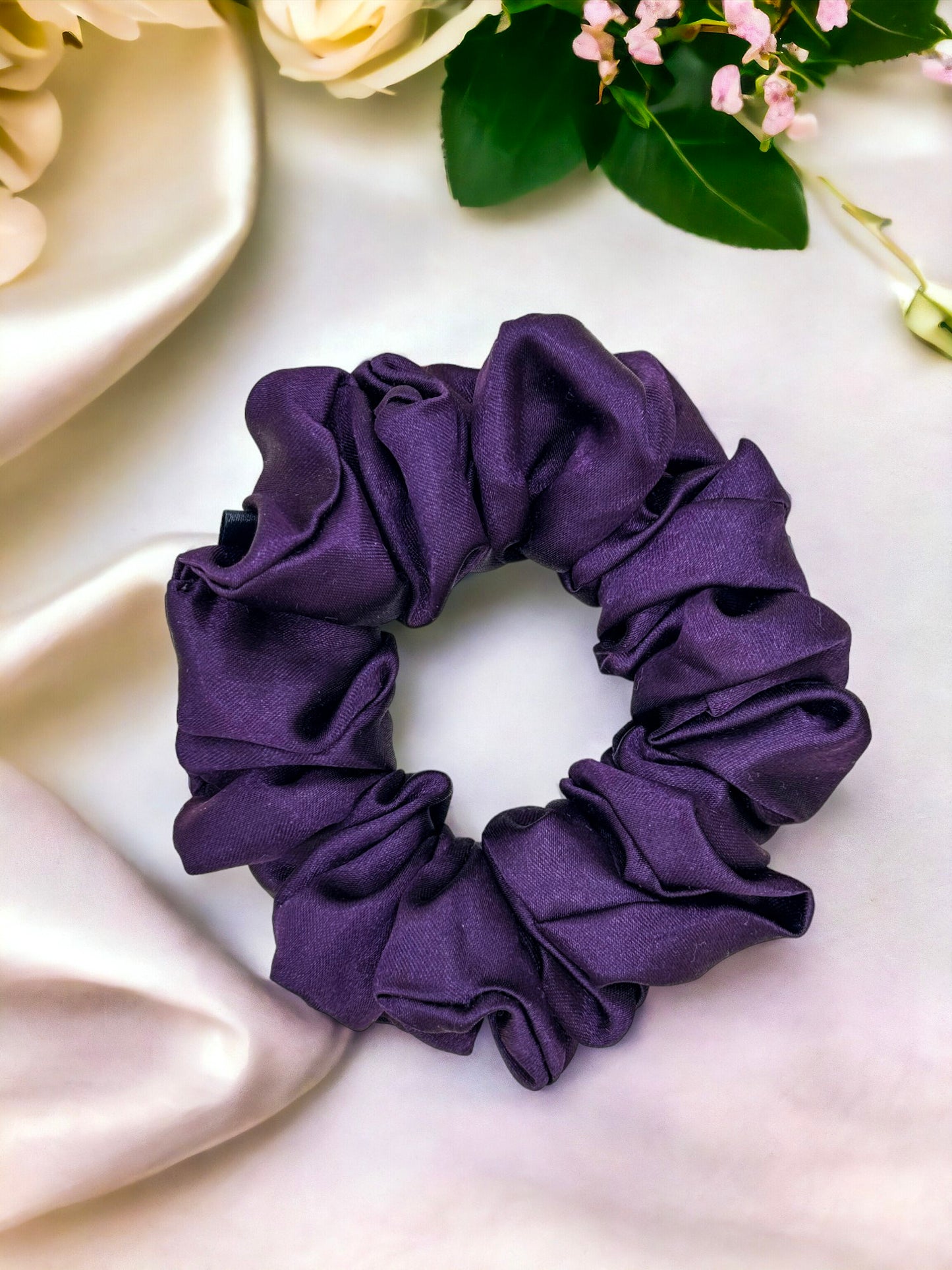 A purple satin Scrunchies-Plain from Bloomnique Outfitters lies on a white surface. Surrounding the handmade scrunchie are white flowers and green leaves, contributing to a soft and elegant aesthetic, perfect for enhancing your hair routine.