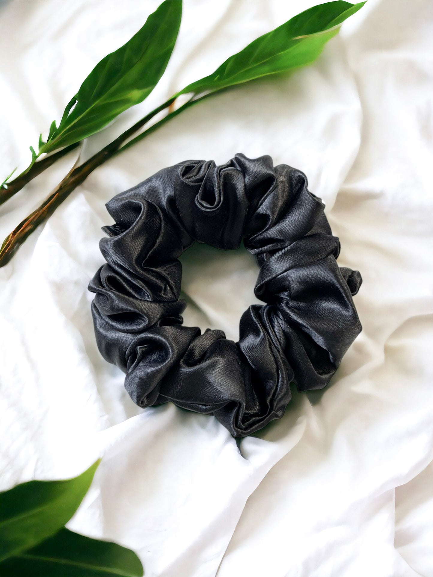 A close-up of a black satin Scrunchies-Plain by Bloomnique Outfitters, perfect for your hair routine, placed on a white fabric background. Two green leaves are partially visible, one on the left and one on the top right, adding a natural element to the composition.