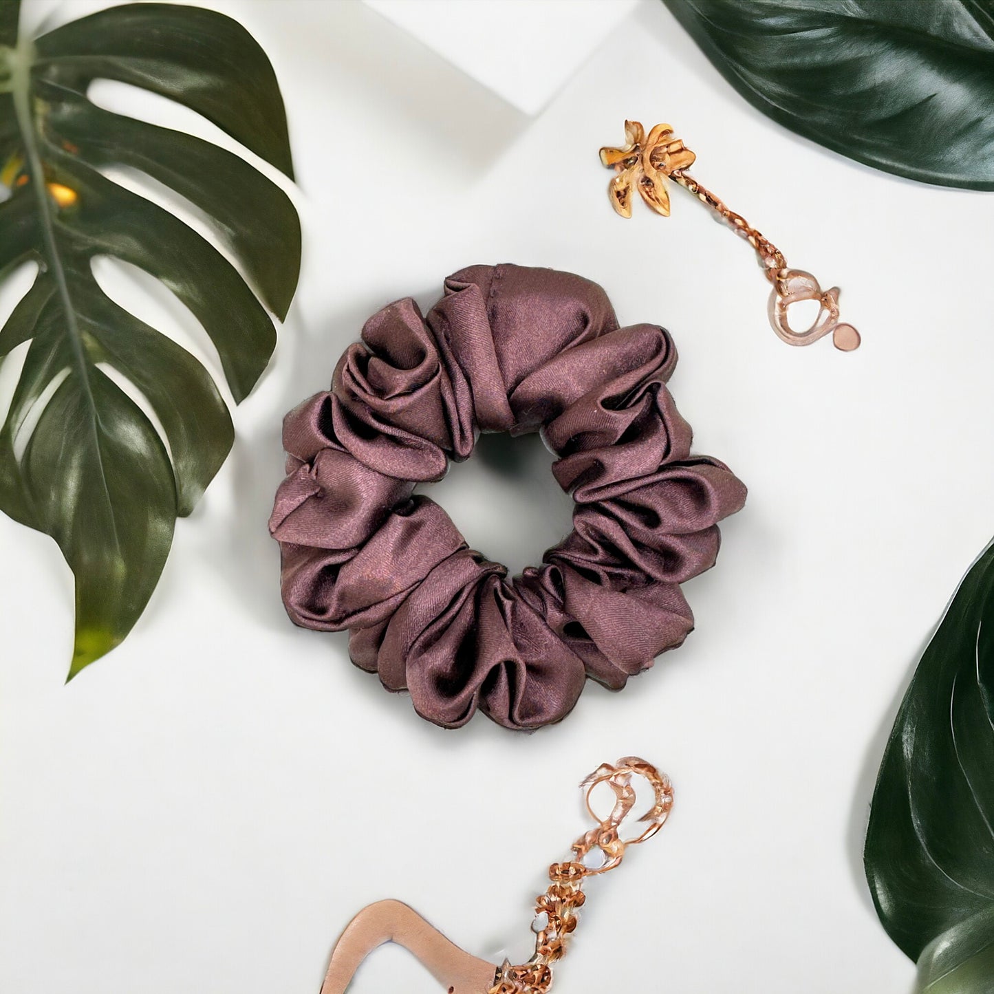 A dark purple satin Scrunchies-Plain by Bloomnique Outfitters lies on a white surface surrounded by green monstera leaves. Nearby, a handmade gold earring with a leaf design and another piece of gold jewelry are also visible, contributing to a nature-inspired aesthetic perfect for your hair routine.