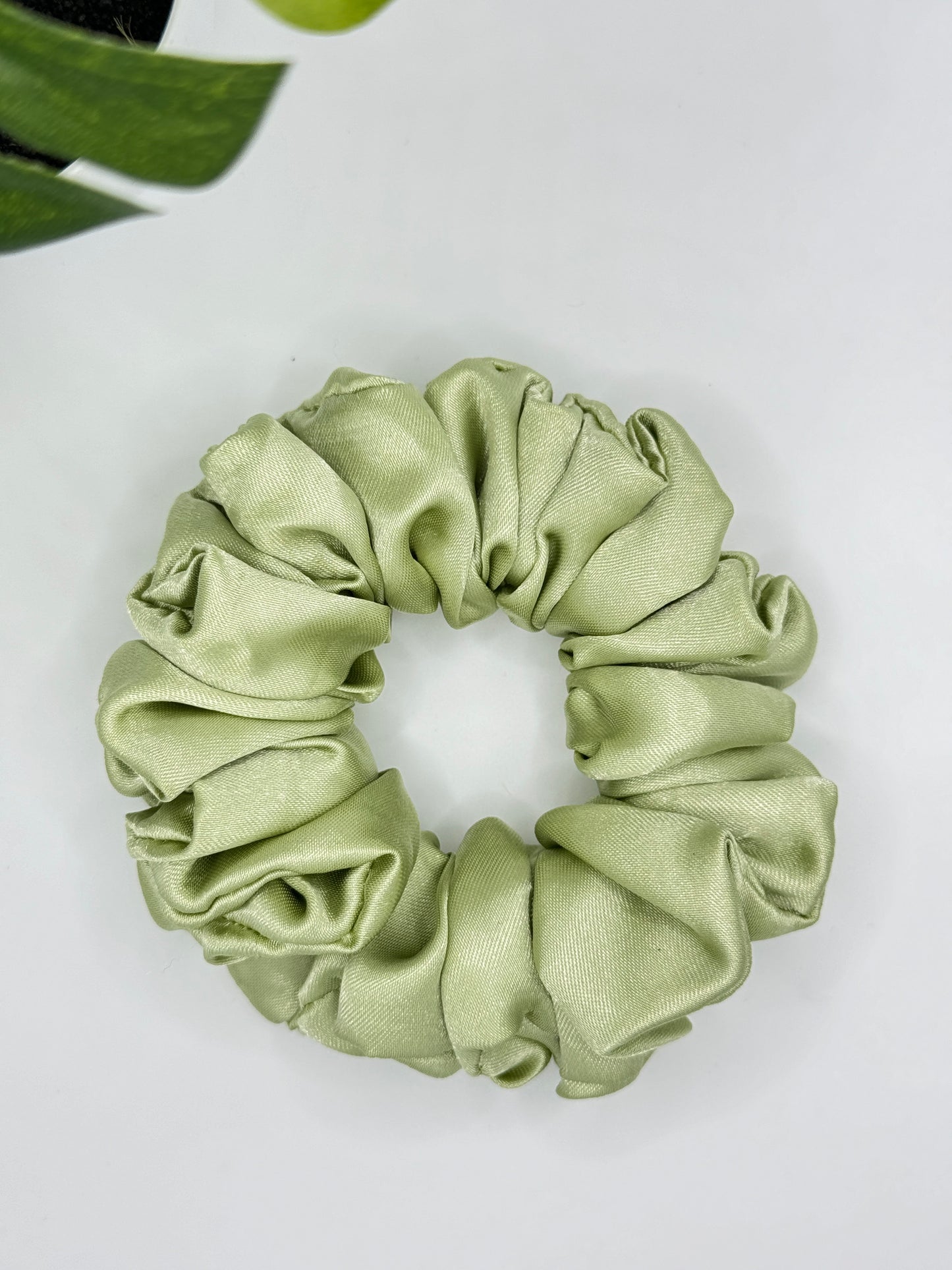A light green, handmade Bloomnique Outfitters Scrunchies-Plain is placed on a plain white background. Partially visible at the top left is a green leaf. The scrunchie has a sheen and a soft, gathered appearance—perfect for elevating any hair routine.