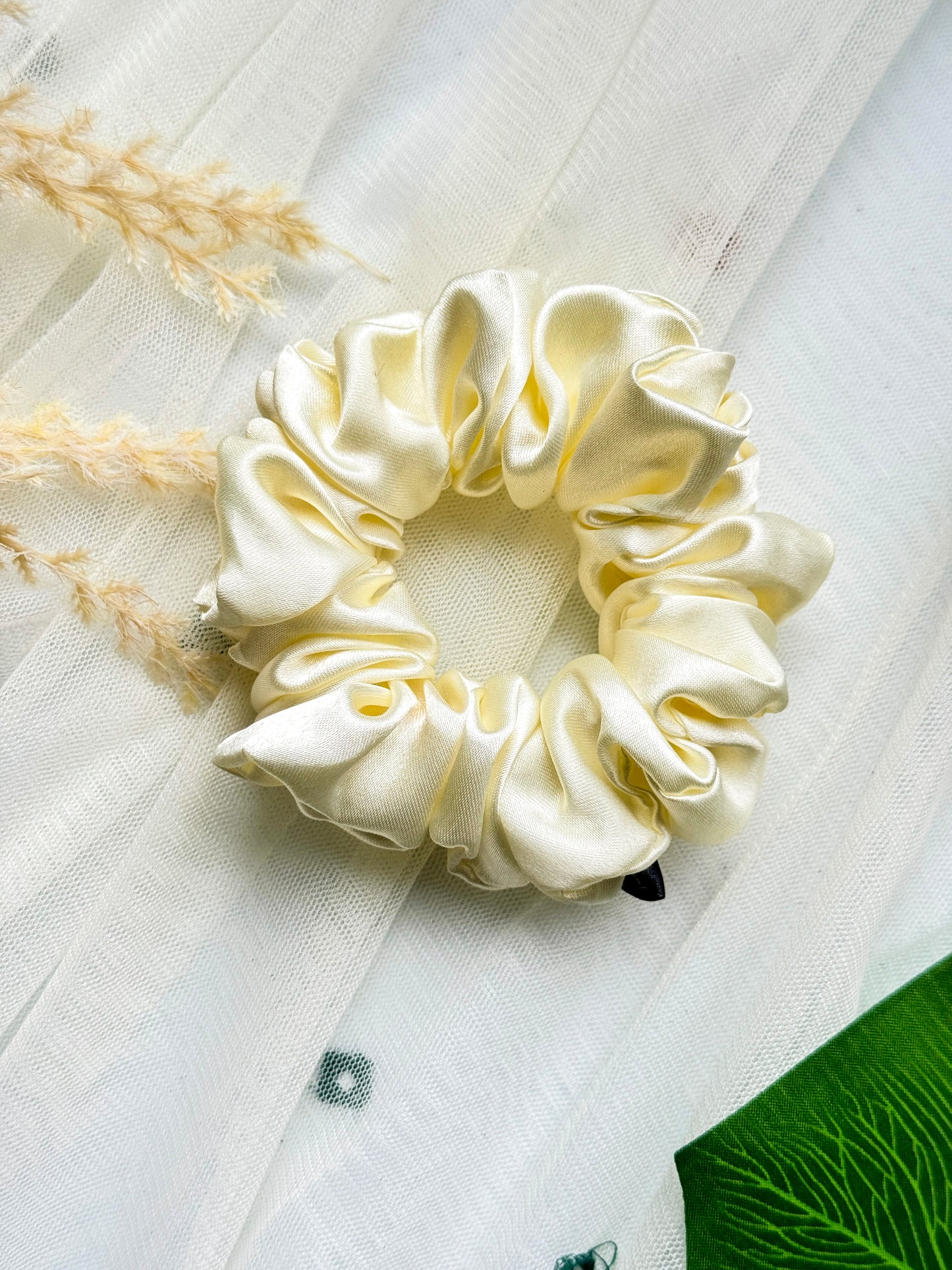 A cream-colored satin scrunchie called Scrunchies-Plain by Bloomnique Outfitters, essential for any hair routine, is placed on a light, sheer fabric surface. Some decorative dried grass and a green leaf can be seen partially in the frame, adding a natural element to the soft and elegant composition.