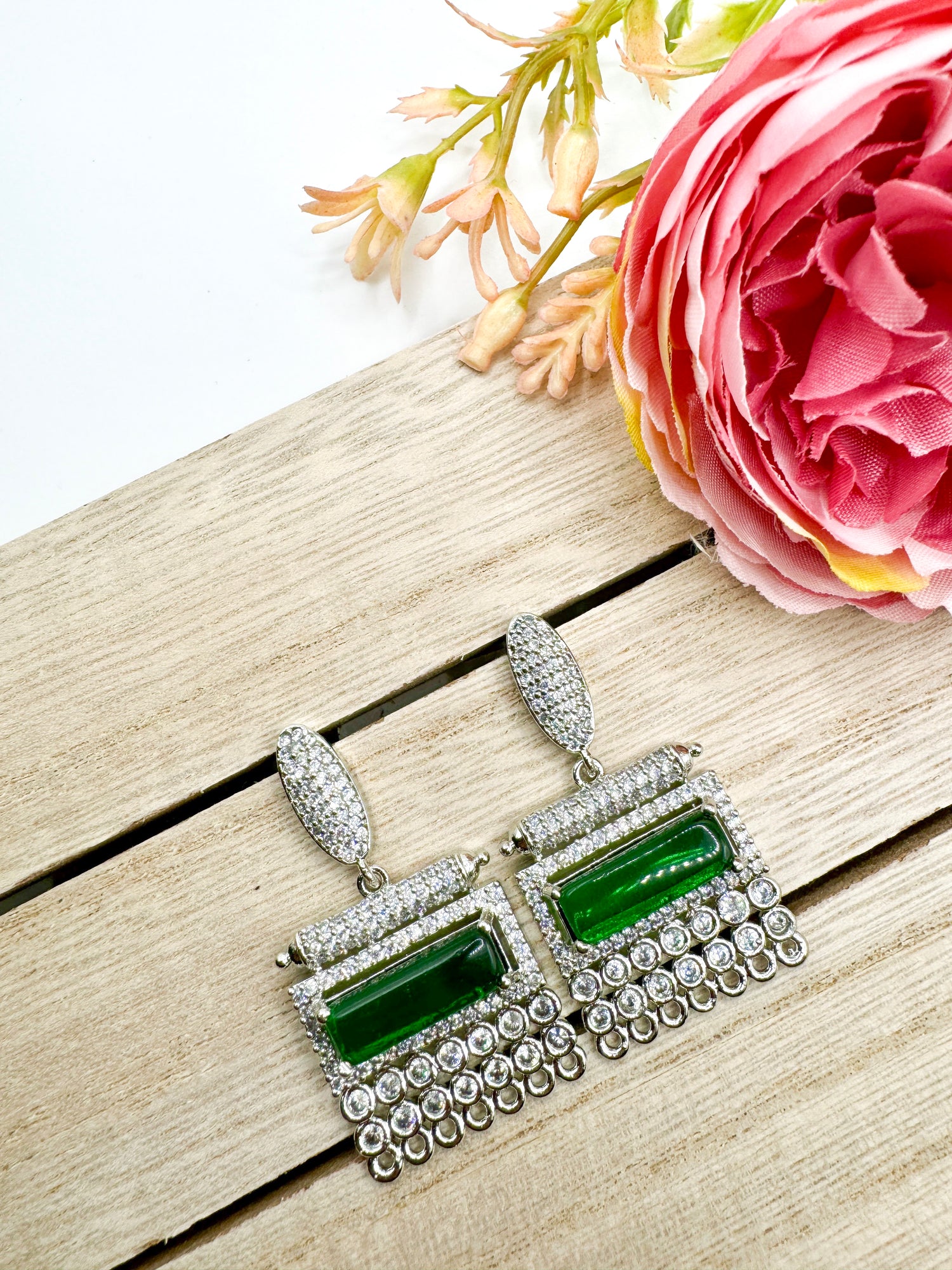 A pair of silver and green earrings lie on a wooden surface near a pink peony flower. The rectangular earrings have emerald-green stones and are adorned with small, sparkling crystals. A few delicate plant sprigs are visible in the background.