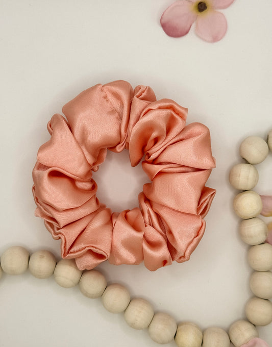 A pink satin Scrunchies-Plain by Bloomnique Outfitters lies on a white surface. To the right and above the scrunchie are partially visible pink flower petals. Below and around the scrunchie are segments of a light wooden bead bracelet or necklace, adding a touch of elegance to your hair routine with handmade accessories.