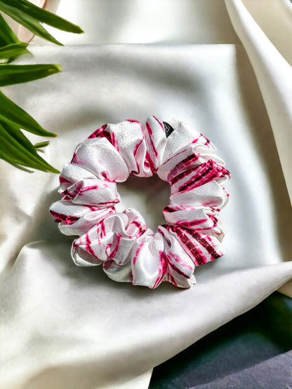 A white and pink striped Scrunchies-Tie Dye by Bloomnique Outfitters lies on a soft, silky white fabric. The handmade satin scrunchie has a slightly shiny texture, giving it an elegant appearance. Some green leaves are partially visible in the top left corner of the image.