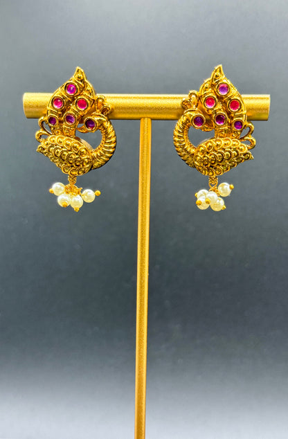 A pair of ornate Peacock Pearl Drop Earrings by Bloomnique Outfitters displayed on a gold stand against a dark gray background. Each earring features intricate designs with embedded red gemstones and small hanging pearl-like decorations at the bottom, exuding an ethnic charm and timeless elegance.