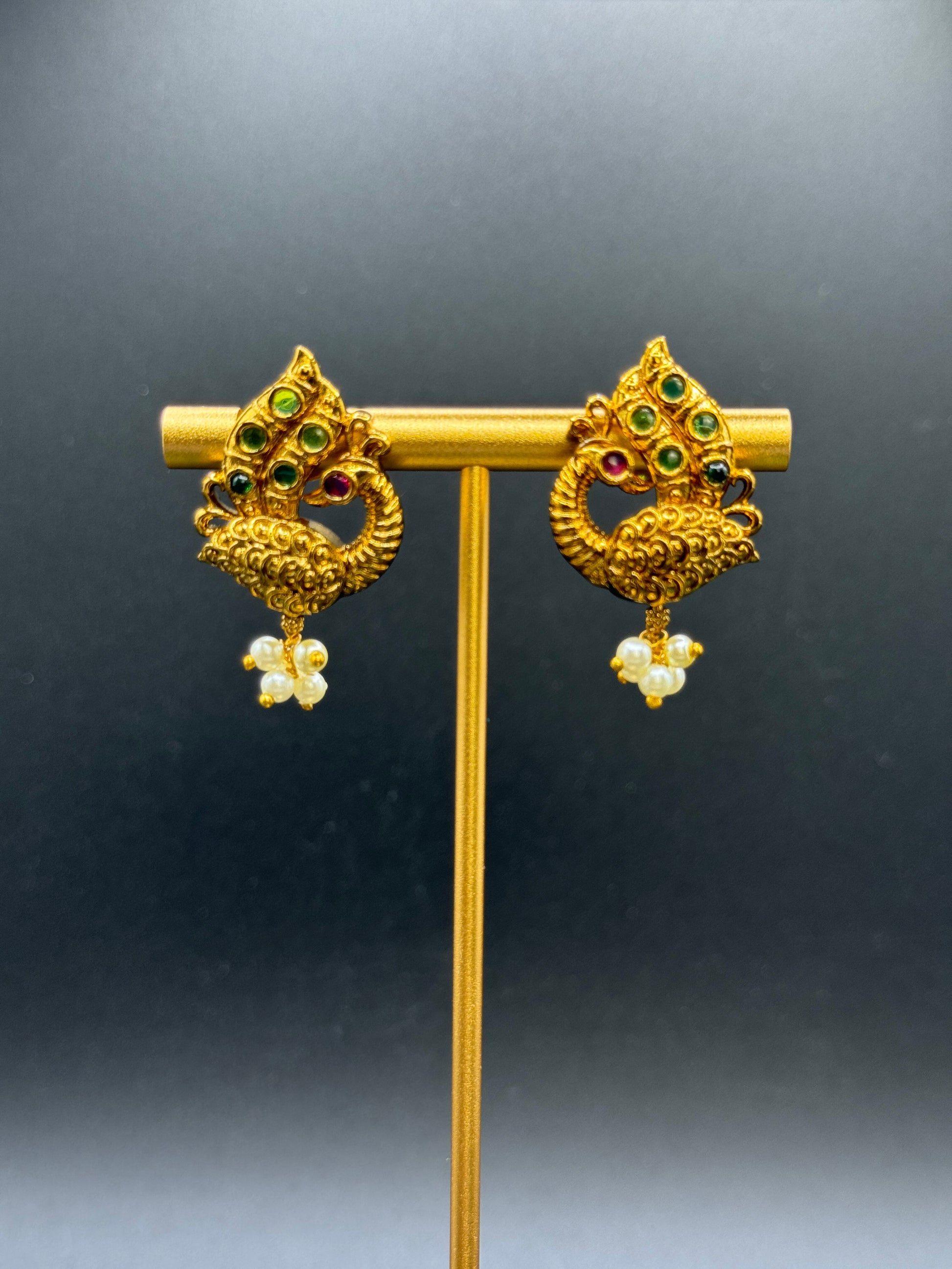 A pair of intricately designed gold-toned Peacock Pearl Drop Earrings from Bloomnique Outfitters with a peacock motif, featuring green, red, and white gemstones. Each earring has small pearl-like beads hanging at the bottom. The earrings exude ethnic charm and are displayed on a stand against a plain, dark background.