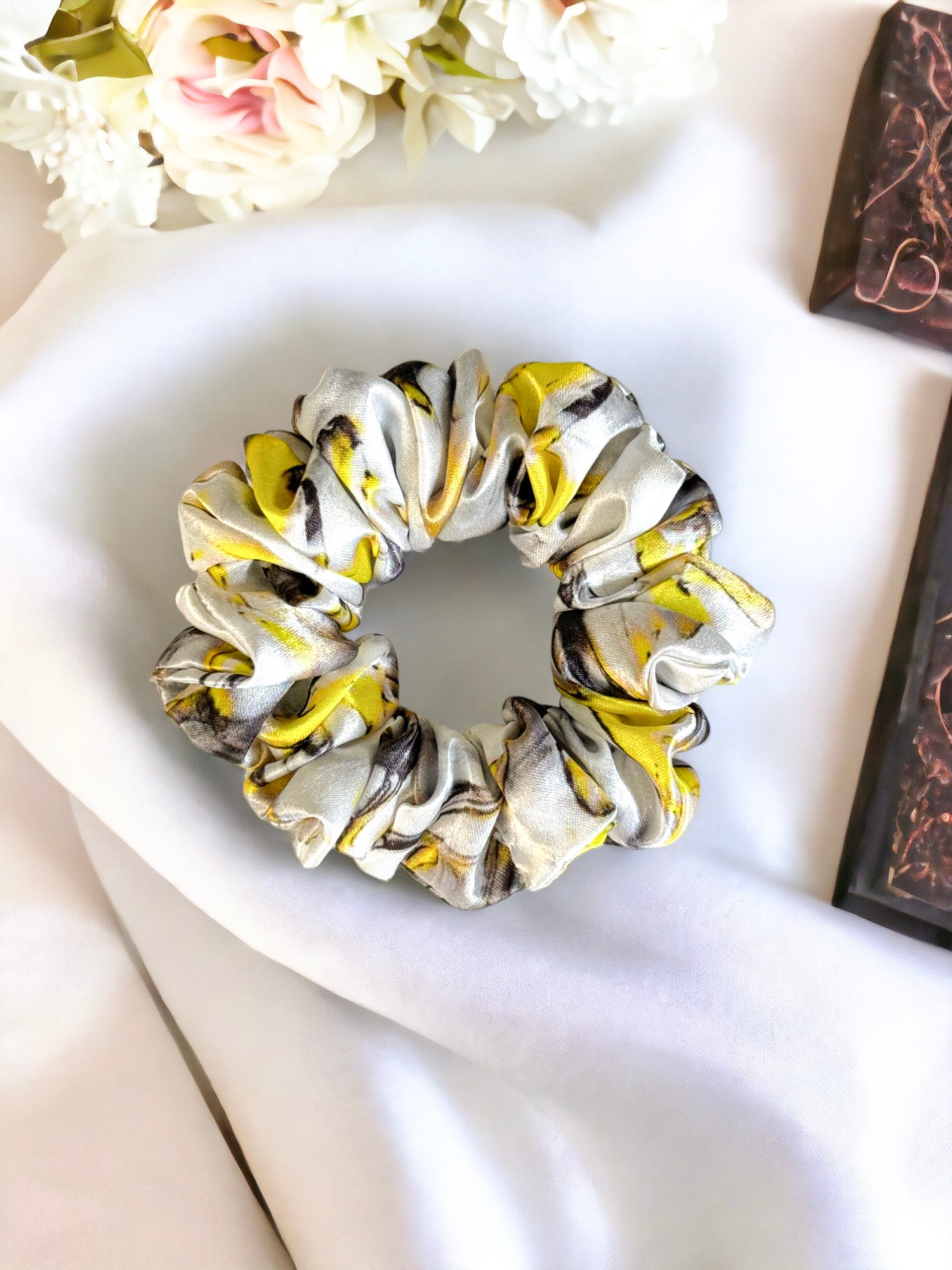 A handmade satin Scrunchies-Tie Dye by Bloomnique Outfitters with a yellow and black pattern rests on a white fabric background. Elegant hair accessories featuring white flowers and a dark brown rectangular object are partially visible in the corners.